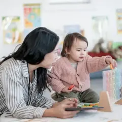 Nauczycielka z tabletem obserwuje dziecko bawiące się klockami. To pokazuje, w jakim wieku idzie się do przedszkola, kiedy nauka i zabawa idą w parze.