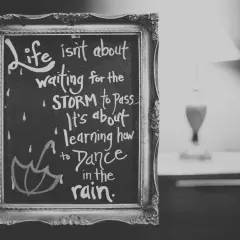 Czarno-biała tablica w ozdobnej ramie z inspirującymi cytatami po angielsku: "Life isn't about waiting for the storm to pass. It's about learning how to dance in the rain.