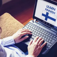 Osoba uczy się języka fińskiego na laptopie, widoczna flaga Finlandii i napis "LEARN FINNISH".