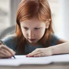 Ruda dziewczynka skupiona na swoich pracach domowych, pisze coś na kartce.