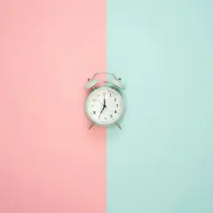 Jasny budzik na tle podzielonym na różowy i turkusowy. Wskazuje godzinę 7:00, więc to poranna godzina am.