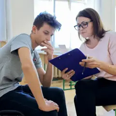 Psycholog szkolny rozmawia z uczniem, który wygląda na zamyślonego. Kobieta trzyma teczkę i długopis.