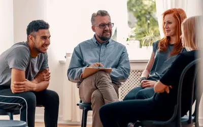 Grupa ludzi na sesji terapeutycznej. Mężczyzna z notatkami prowadzi rozmowę, a uczestnicy słuchają. Może uczą się języka francuskiego?