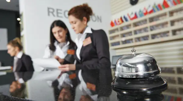 Recepcjonistki w hotelu omawiają dokumenty. Dzwonek hotelowy na pierwszym planie. Zwroty po angielsku w hotelu na recepcji są kluczowe.