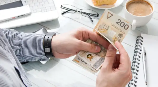 Ręce liczą polskie banknoty 200 zł. Obok laptop, kawa, rogalik i notatnik. To obrazek pokazujący klasa średnia zarobki.