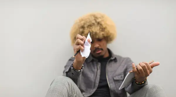 Mężczyzna z afro włosami siedzi, trzymając ołówek i zmiętą kartkę papieru.