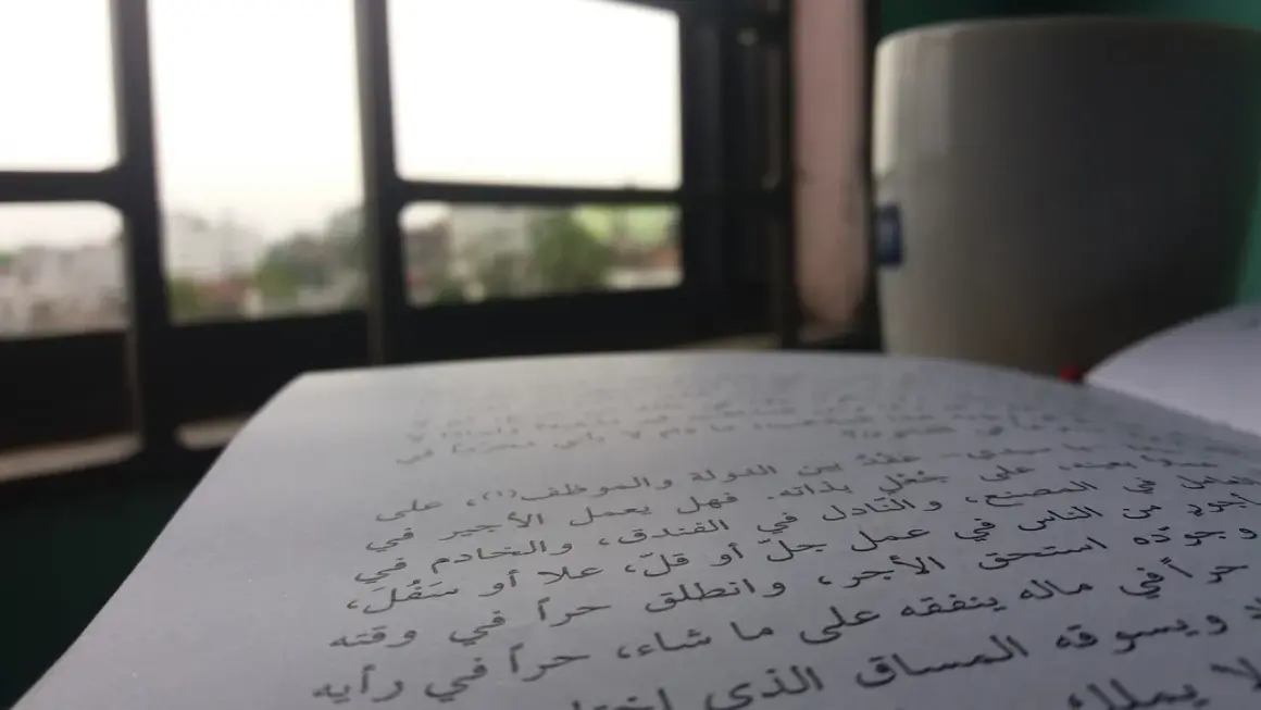 Otwarta księga z tekstem w języku arabskim, obok kubek i widok przez okno.