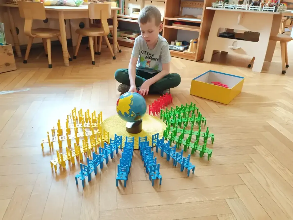 Chłopiec bawi się klockami w kształcie krzesełek, tworząc okręgi wokół globusa. To nauka przez zabawę w stylu metoda montessori.