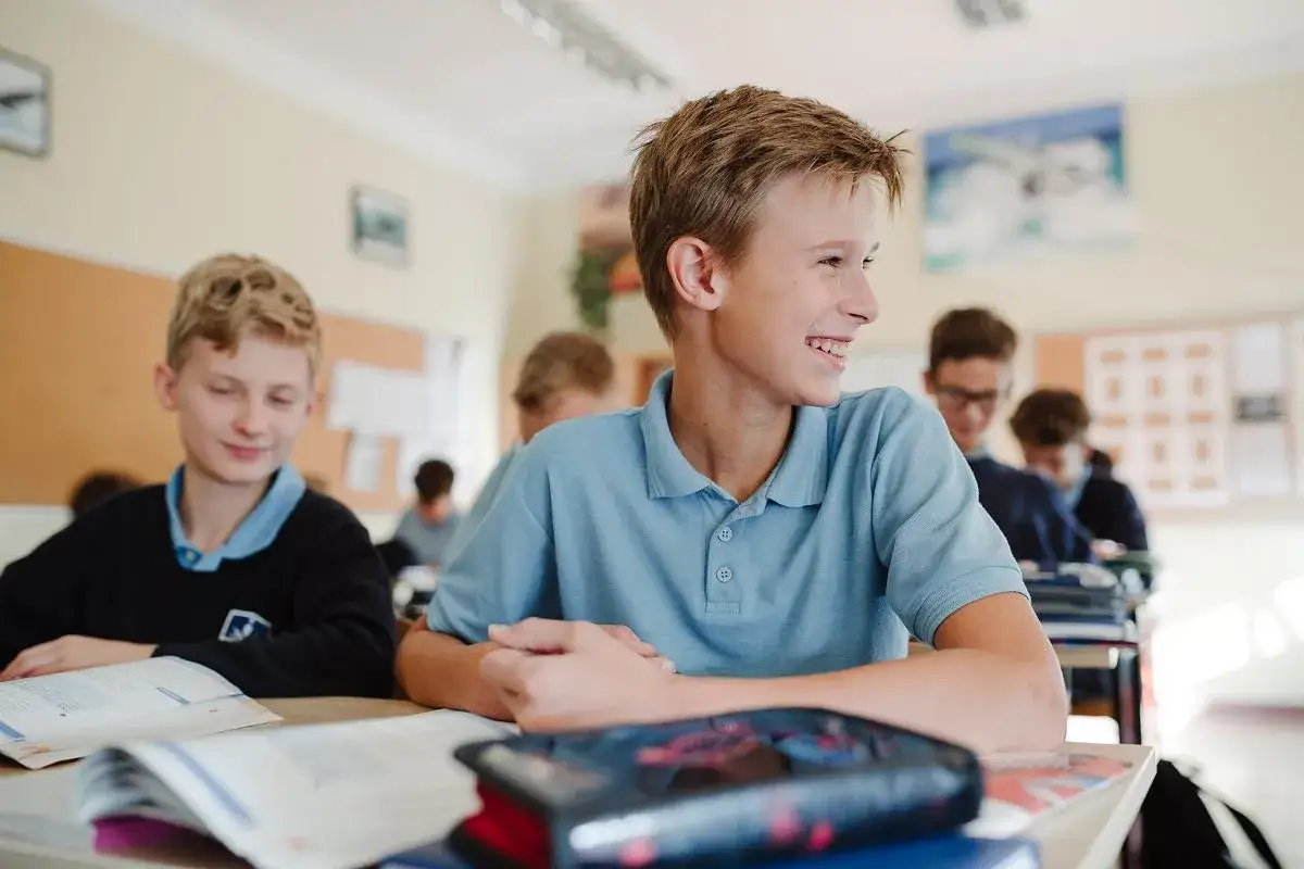 Uśmiechnięty chłopiec na lekcji. Czy jest obowiązek nauki do 18 roku życia? W klasie siedzą uczniowie, każdy pochłonięty swoimi zadaniami.