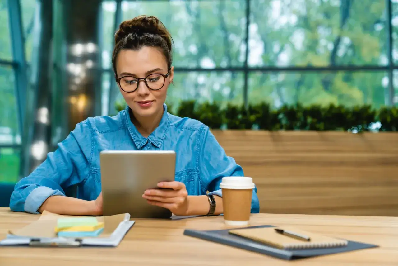 Kobieta w okularach przegląda tablet, szukając informacji o apple zniżka studencka. Obok stoi kubek kawy.