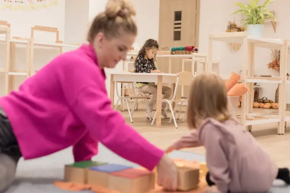Nauczycielka wspiera dzieci w nauce metodą Montessori. Dzieci bawią się klockami i rysują przy stoliku.